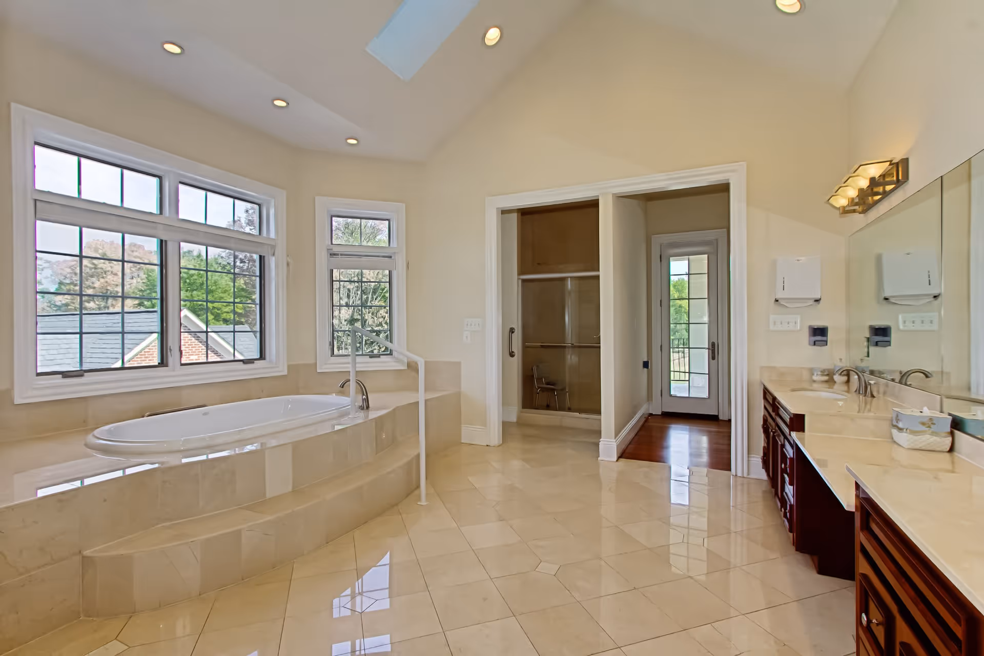 Spacious bathroom with a large built-in bathtub surrounded by beige tiles, two large windows with grid patterns letting in natural light, a walk-in shower with a glass door, a door leading outside, and a long vanity with two sinks, mirrors, and wall-mounted soap and paper towel dispensers.
