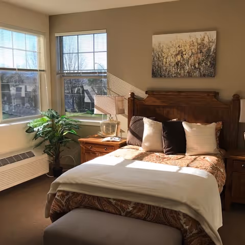 Sunlit bedroom with a wooden bed and headboard, nightstands and lamps, a potted plant, and large windows.