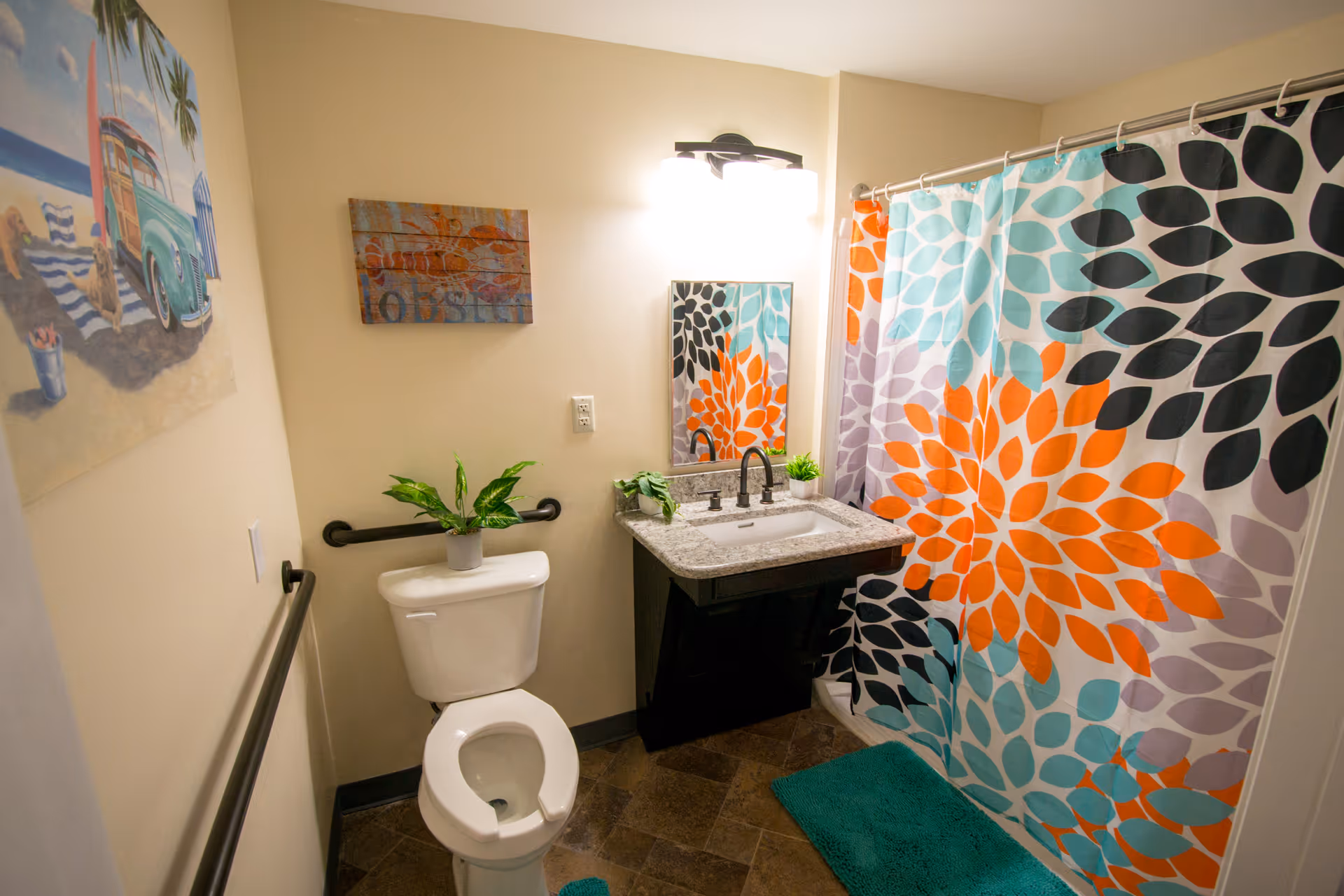 A bathroom with a white toilet and a granite countertop sink with black fixtures. The shower has a colorful curtain with large floral patterns in orange, black, teal, and purple. There are two green plants on the sink and toilet tank. The walls are beige with two pieces of wall art, one depicting a beach scene with a vintage car and the other a rustic wooden sign. The floor has brown tiles and there are teal bath mats on the floor. Black grab bars are installed near the toilet and along the wall.