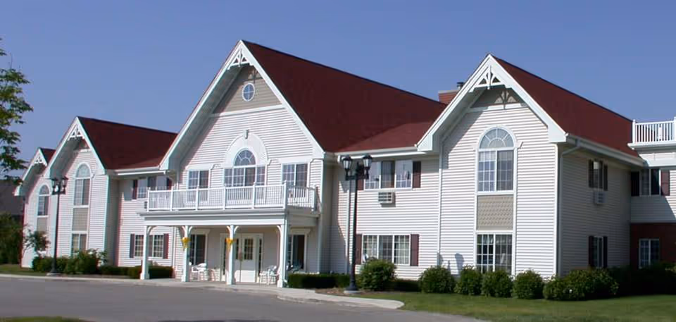 Exterior view of a large two-story senior living facility building with white siding and a red roof under a clear blue sky. The building features multiple windows, a balcony, and a covered entrance with chairs outside. There are bushes and a lamp post near the entrance.