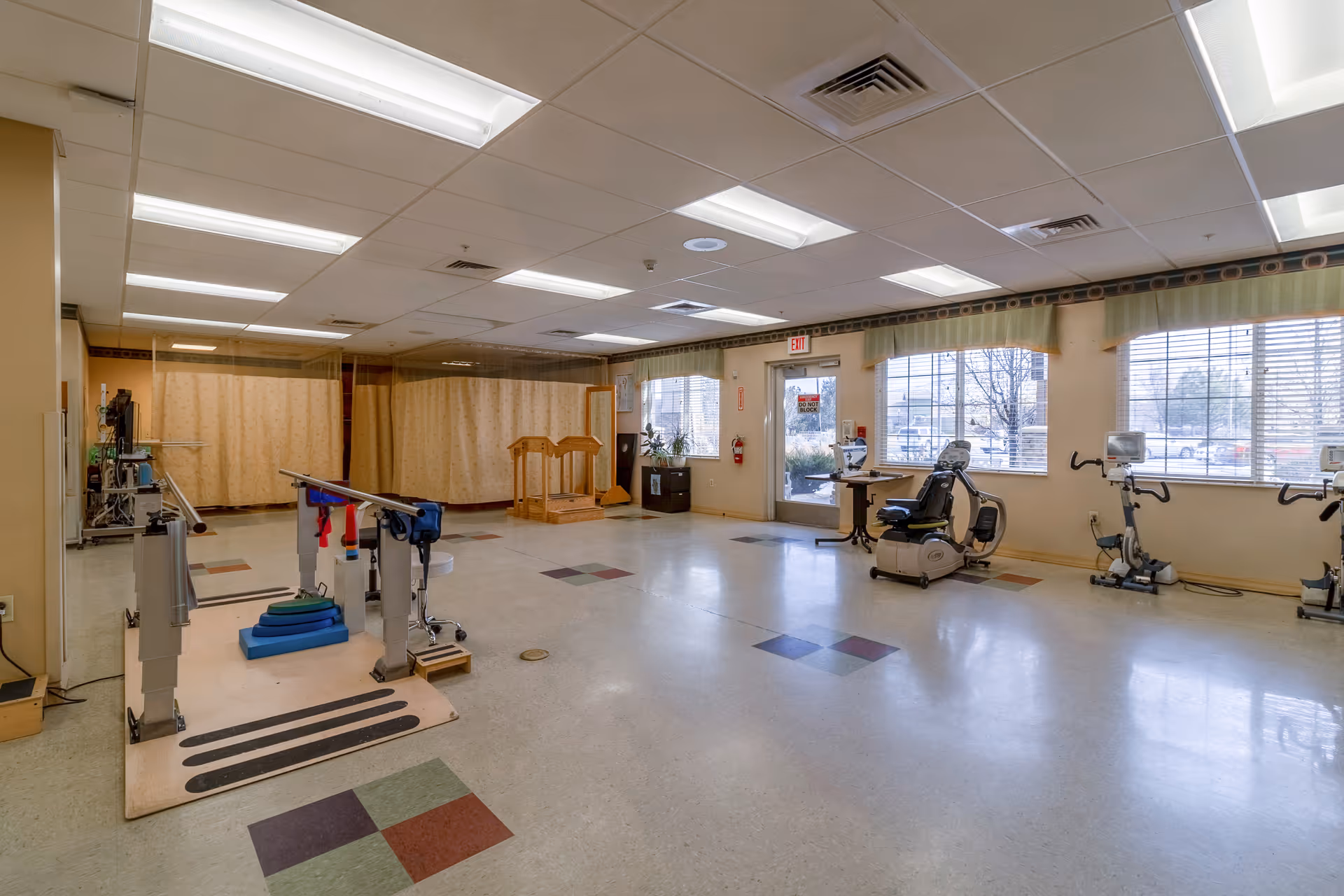 A spacious rehabilitation room with parallel bars for walking exercises, exercise bikes near large windows, and a wooden step platform. The room has tiled floors with colored square patterns, beige walls, and fluorescent ceiling lights. There are large windows letting in natural light and an exit door with a 'Do Not Block' sign.