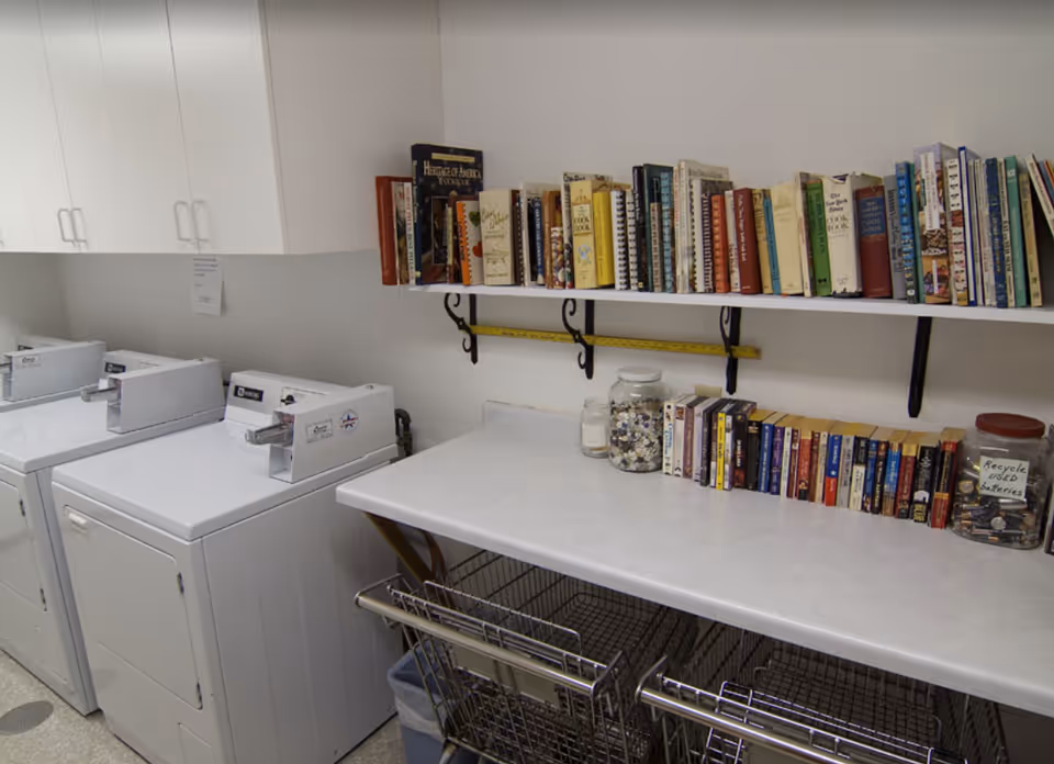 Laundry room with three white washing machines and dryers on the left side, white cabinets above them, a countertop on the right with two jars filled with buttons and a jar labeled 'Recycle used batteries', and a shelf above the counter holding a variety of books.