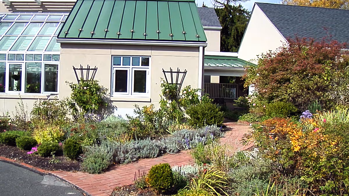 Front exterior of a light-colored building with a green metal roof, a glass sunroom, and a brick walkway through landscaped gardens.