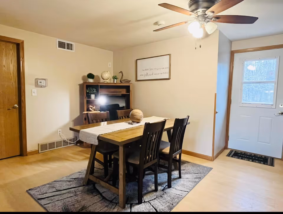 Wooden dining table with four chairs on a rug beneath a ceiling fan, next to a white entry door and a small shelving unit.