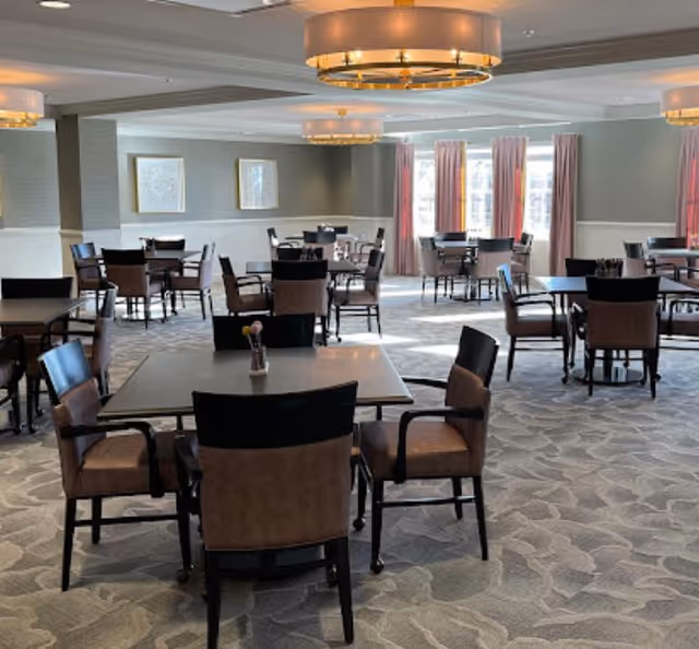 Spacious dining room with multiple square tables and upholstered chairs under modern chandeliers and windows with pink curtains.