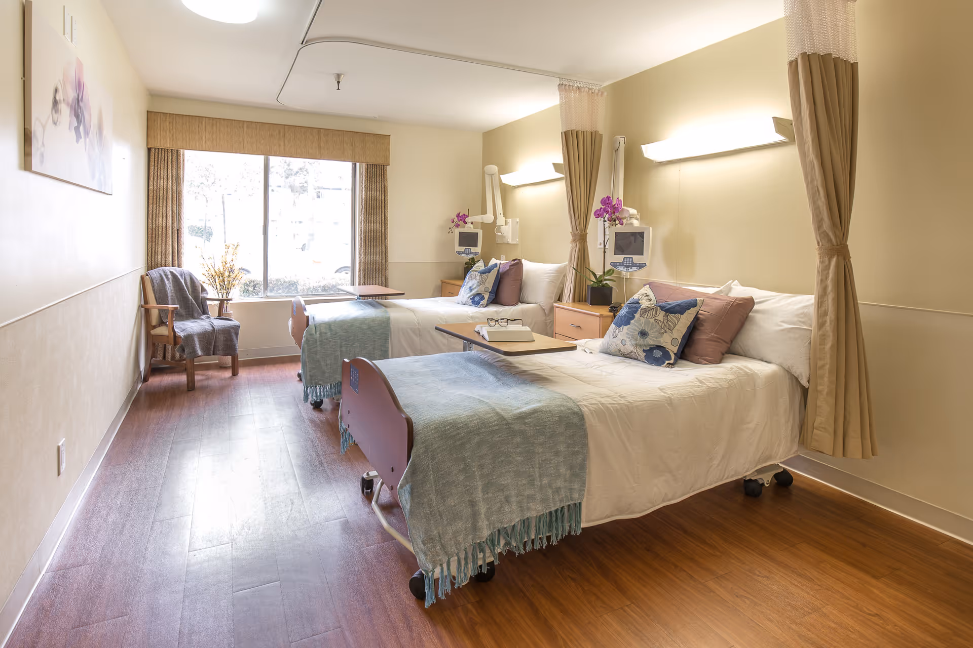 A bright and clean convalescent center room with two hospital beds, each made with white linens, blue throws, and decorative pillows. Between the beds is a wooden nightstand with a purple orchid plant. Each bed has a mounted medical monitor and an adjustable overbed table. The room has wood flooring, a large window with beige curtains, and a wooden chair with a gray blanket draped over it. Soft wall lights illuminate the space.