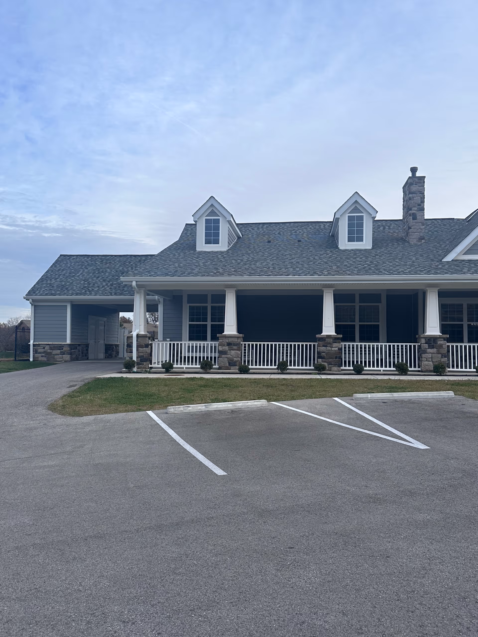 Front exterior view of a single-story building with a gray roof, two dormer windows, stone pillars supporting a covered porch with white railings, and a parking area in front with marked spaces.