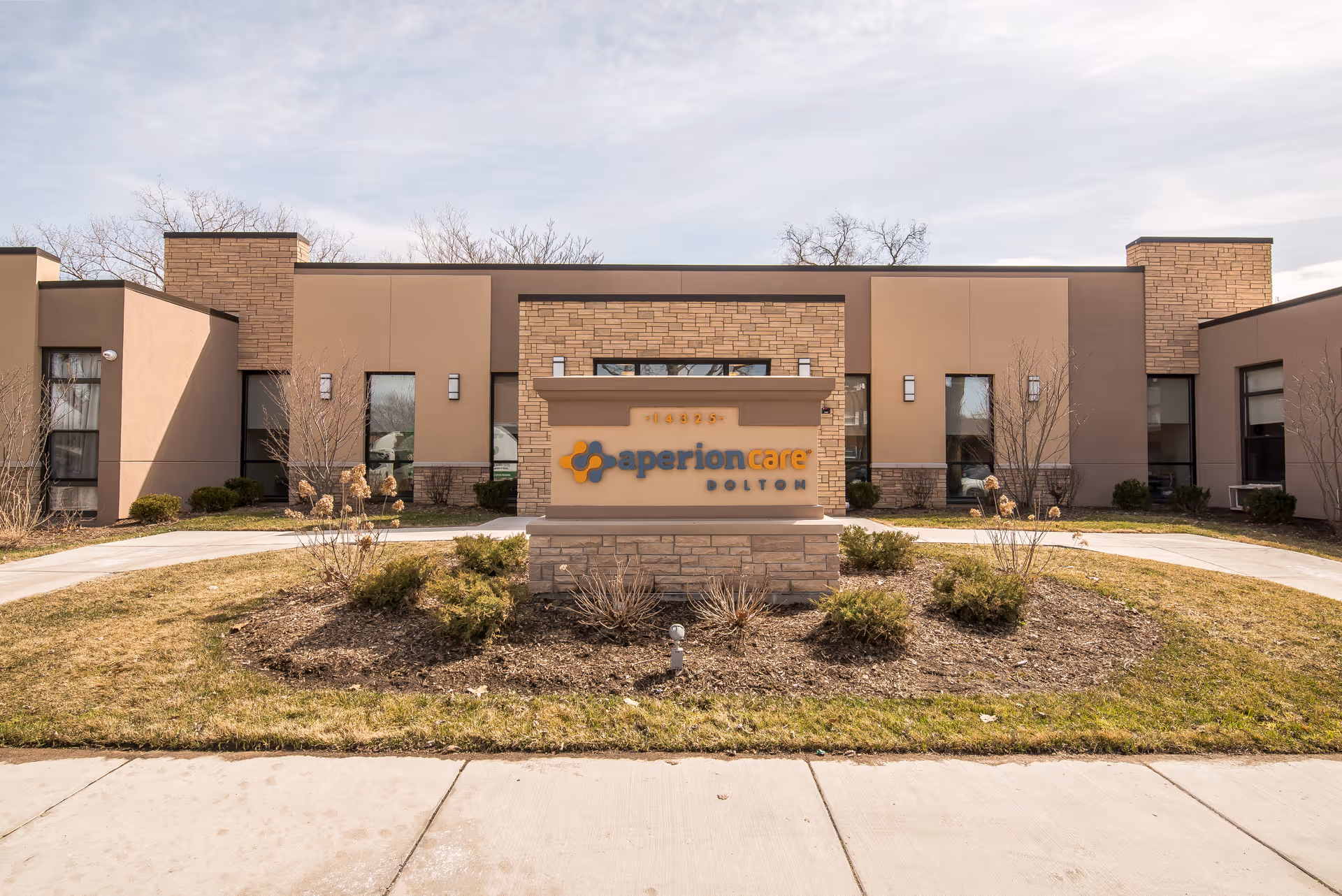 Front exterior view of Aperion Care Dolton building with a stone and beige facade, a sign displaying the facility name in front, surrounded by landscaped bushes and a curved walkway.