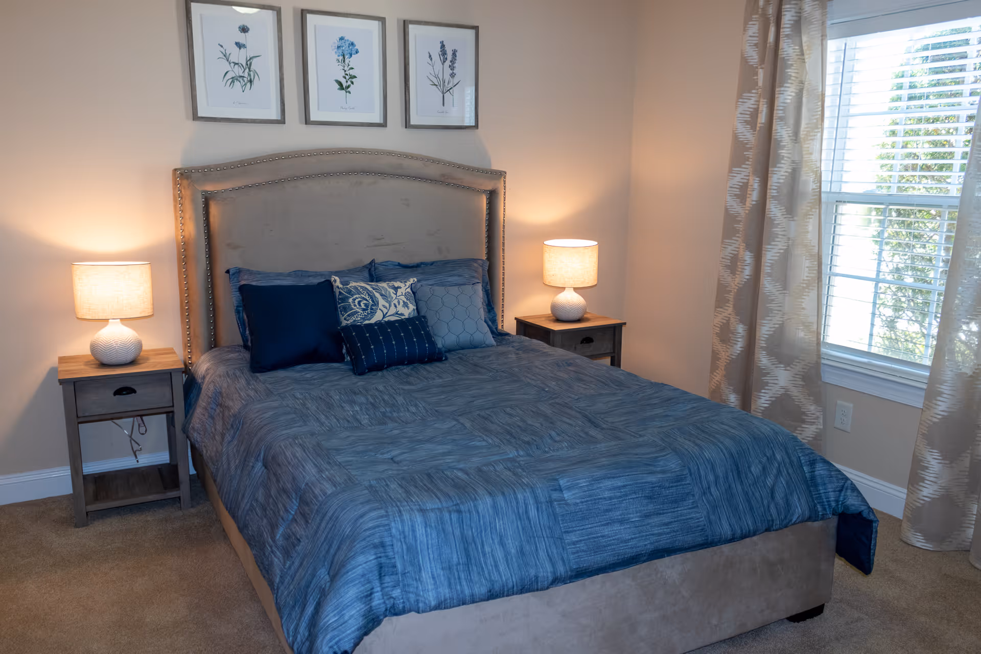 A cozy bedroom with a beige upholstered headboard and a bed dressed in blue bedding and multiple decorative pillows. There are two wooden nightstands on either side of the bed, each with a lit table lamp. Above the headboard, three framed botanical prints are hung on the wall. A window with patterned curtains allows natural light into the room.