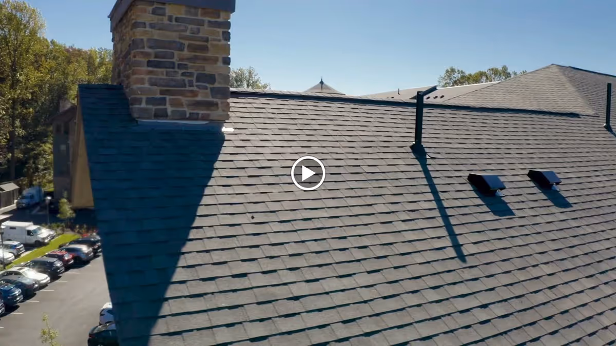 Aerial view of a sloped shingled roof with a stone chimney and vents, with a parking lot and trees visible in the background.