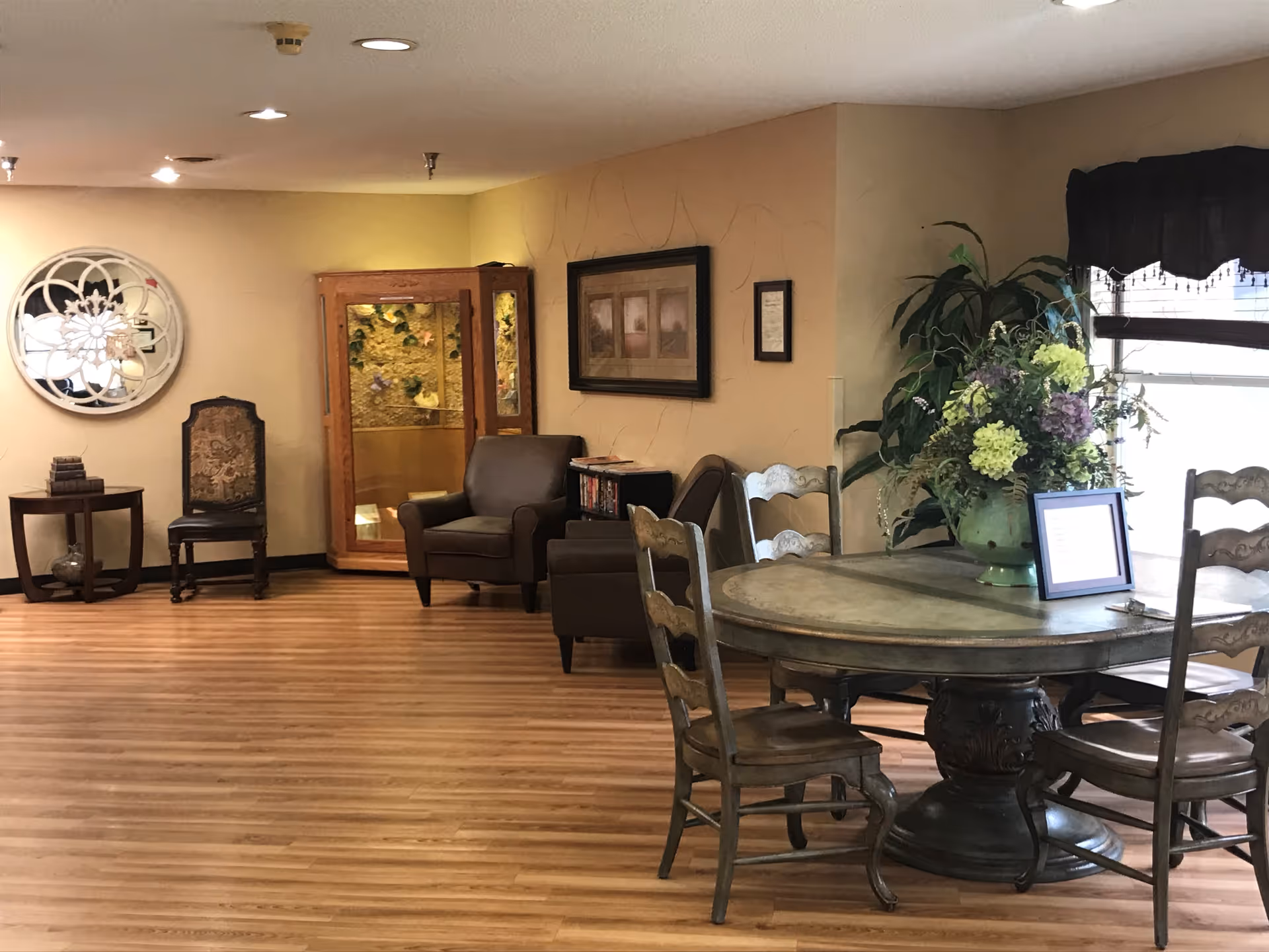 A cozy interior common area with wooden flooring, a round wooden table with four chairs, a large floral arrangement on the table, two armchairs, a decorative wall mirror, framed artwork, and a window with a dark valance allowing natural light.
