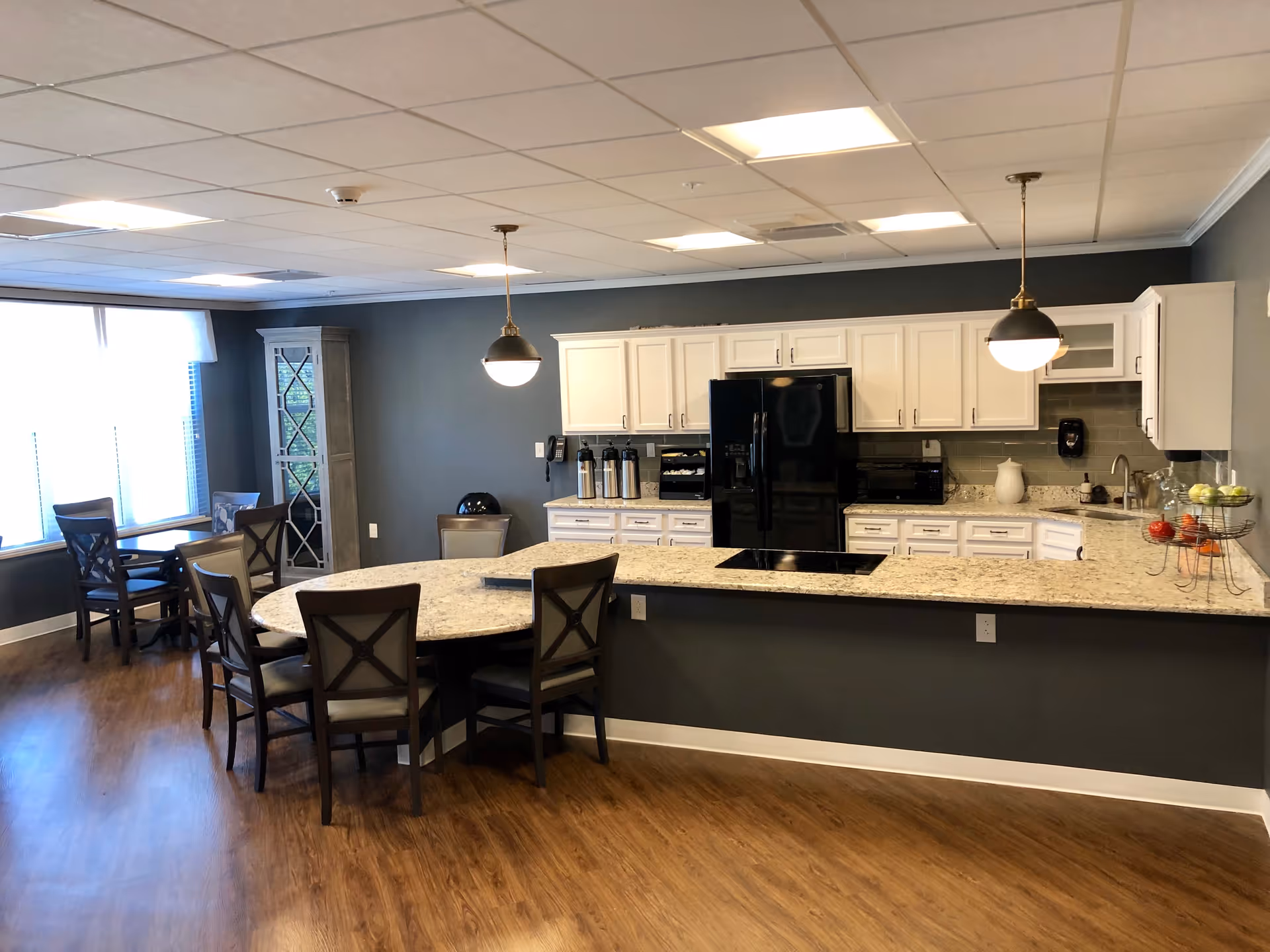 A modern kitchen and dining area with white cabinets, a black refrigerator, granite countertops, and wooden chairs around a curved dining table. The room has wooden flooring, pendant lights hanging from the ceiling, and a large window with blinds letting in natural light.