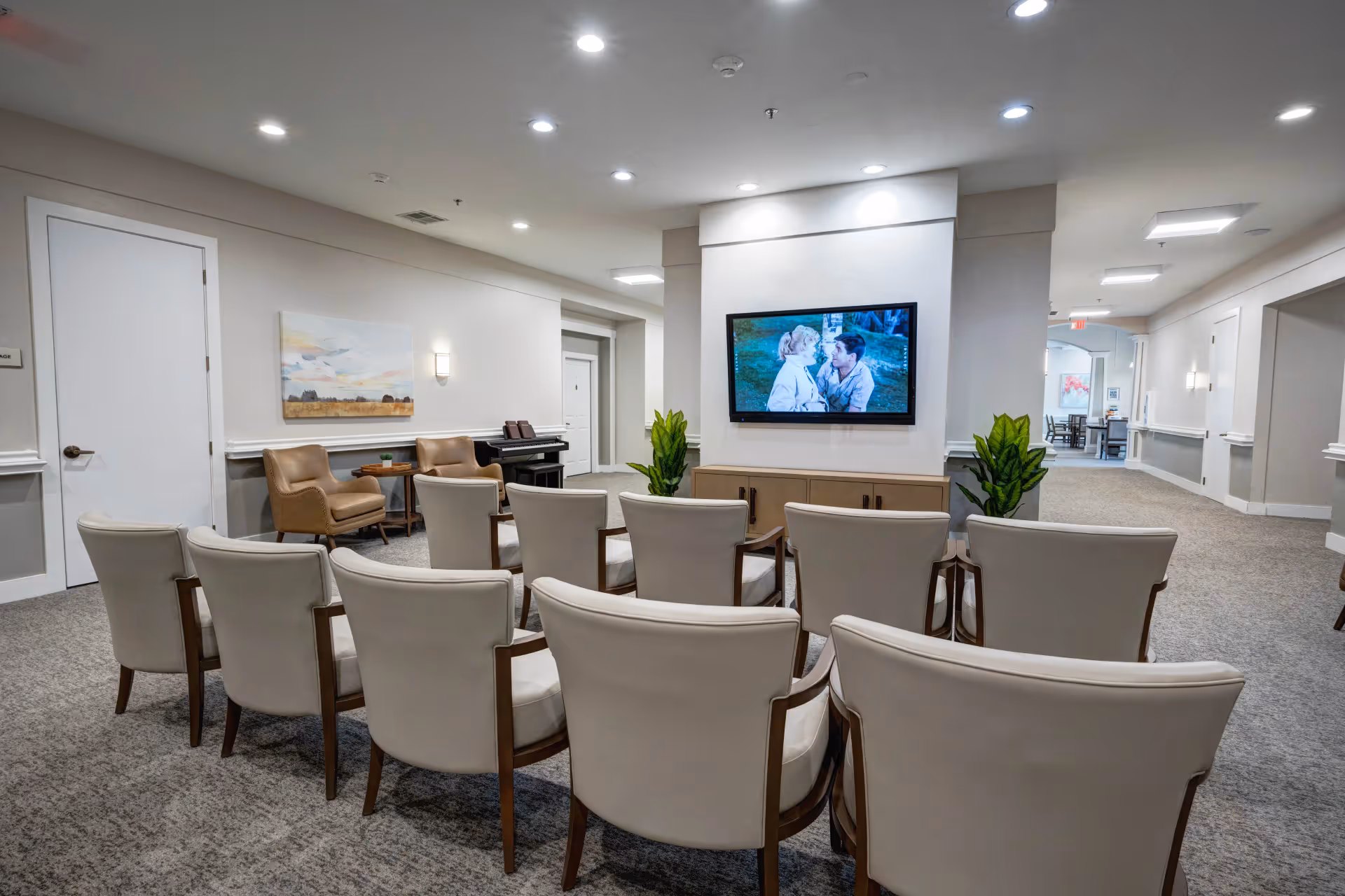 A common area in an assisted living facility with two rows of beige chairs facing a wall-mounted television. The TV is showing a scene with two people. The room has neutral-colored walls, carpeted floors, a couple of potted plants, and additional seating with two tan armchairs and a small table near a piano. The hallway extends to the right with more doors and lighting fixtures.