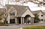 Front exterior of a multi-story senior living facility with a covered entrance and columns, trees, and a driveway.