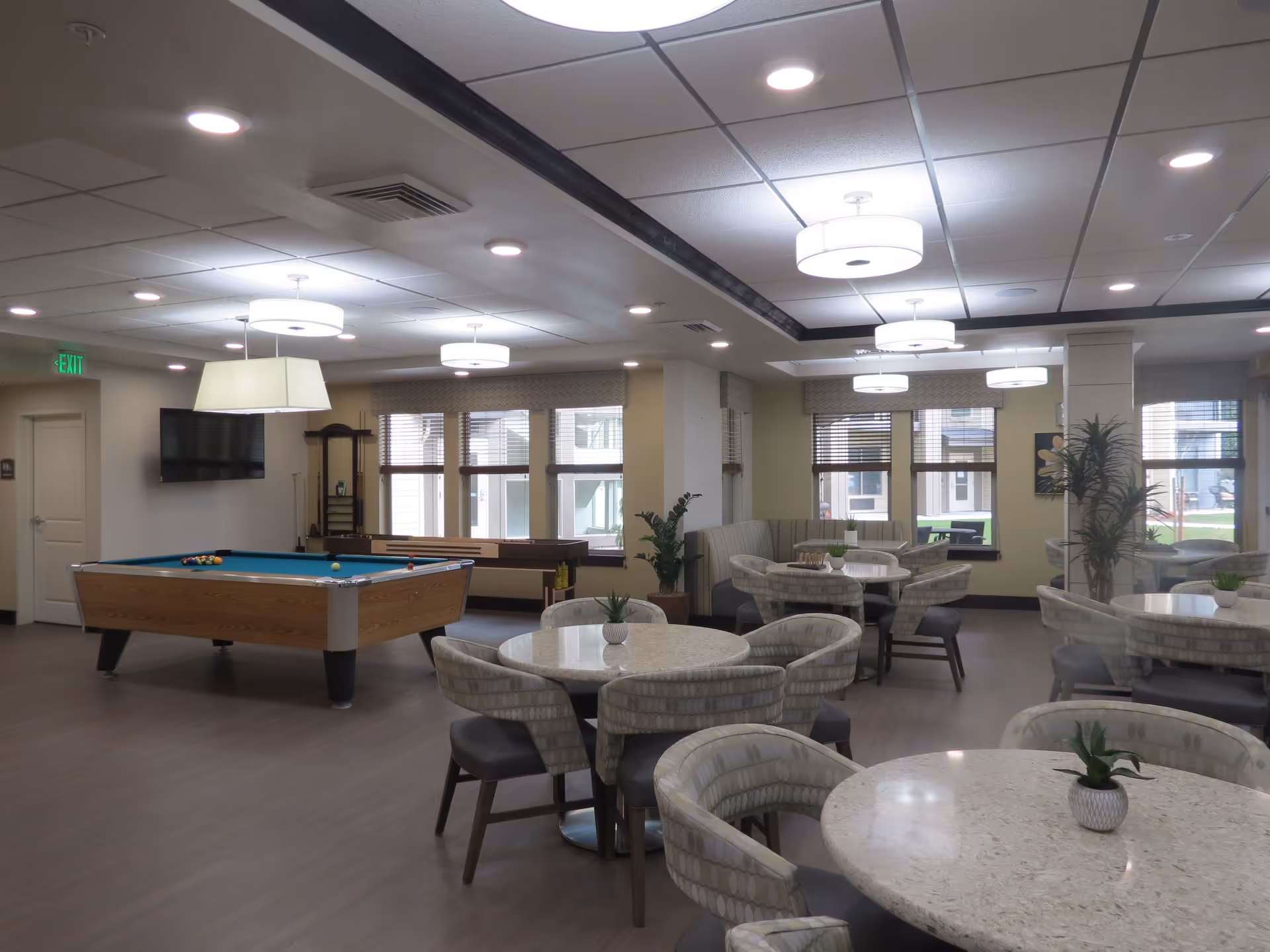 A bright communal activity room with a pool table, round dining tables and upholstered chairs under pendant lights.