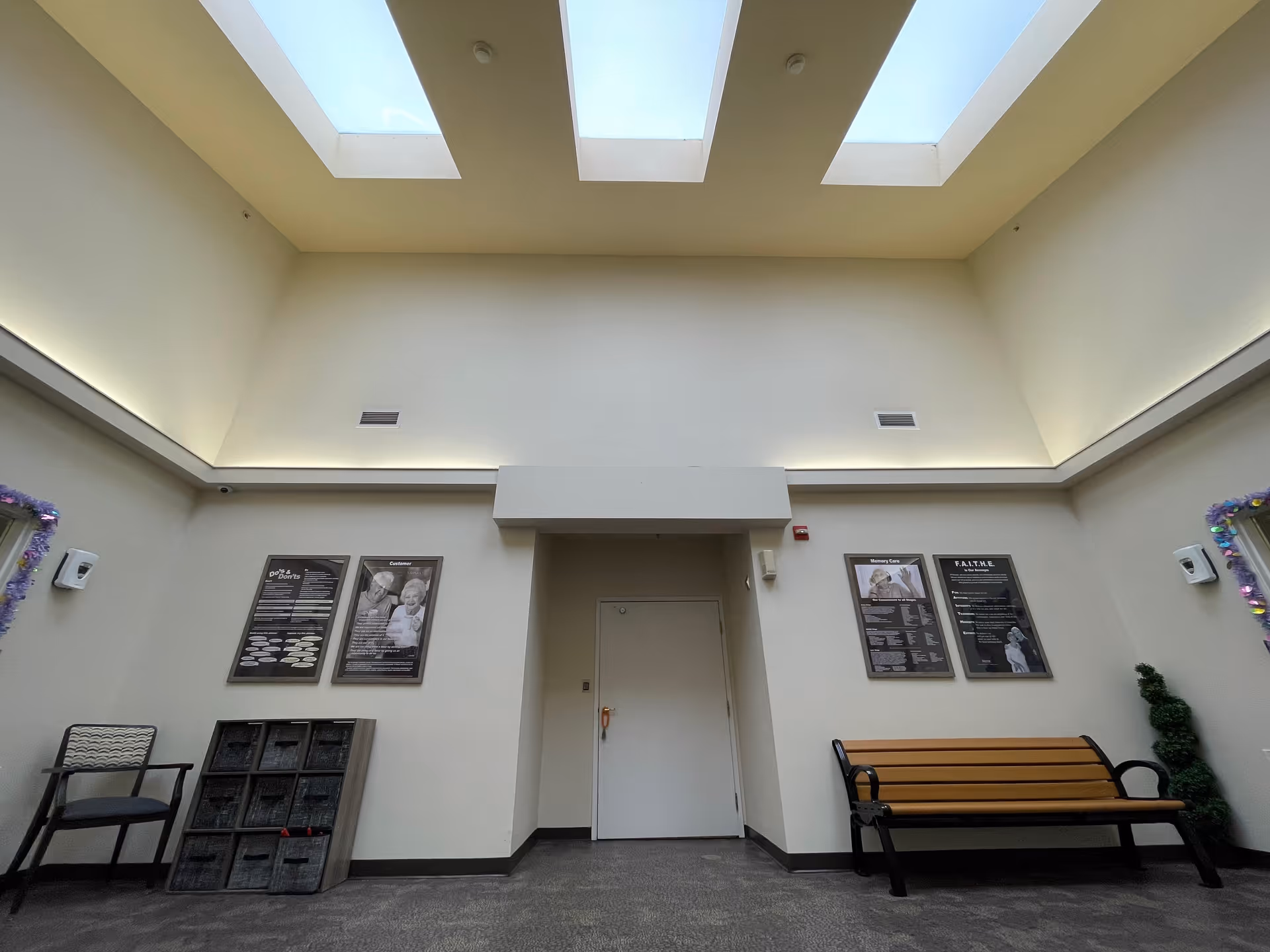 Interior view of a senior living facility lobby or common area with a high ceiling featuring three skylights. The room has light-colored walls with informational posters mounted on them. There is a wooden bench with black metal armrests on the right side and a single chair on the left side. A small decorative plant is placed near the bench.