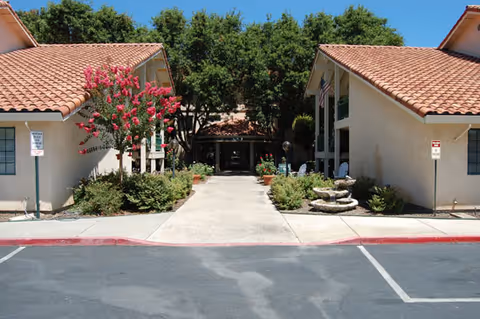 Entrance walkway to a senior living facility with beige stucco buildings on either side, red tile roofs, landscaped bushes and a flowering tree, leading to a shaded entrance surrounded by large trees.