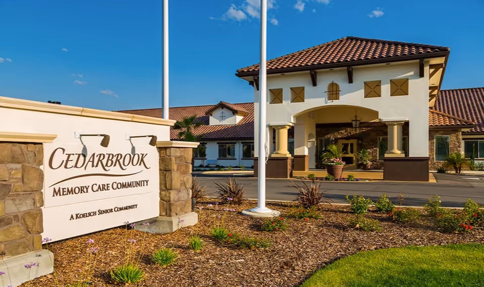 Exterior view of Cedarbrook Memory Care Community building with a stone and stucco sign in the foreground and the main entrance with a covered driveway in the background under a clear blue sky.