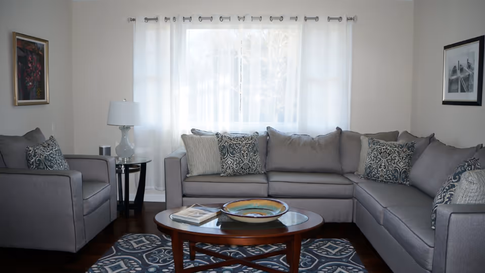 A cozy living room with a large gray sectional sofa and a matching armchair, both adorned with patterned throw pillows. A wooden coffee table with a decorative bowl and a book sits on a blue and white patterned rug. A glass side table with a lamp is next to the armchair. The room has light-colored walls, two framed pictures, and a large window with sheer white curtains letting in natural light.