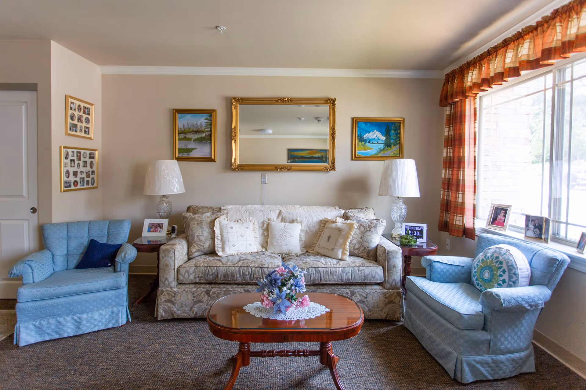 A cozy living room with a floral-patterned sofa adorned with multiple cushions, flanked by two blue upholstered armchairs. A wooden coffee table with a floral centerpiece sits in front of the sofa. Two table lamps with white shades are placed on side tables beside the sofa. The walls are decorated with framed paintings and a large ornate mirror. A window with orange plaid curtains allows natural light to fill the room.
