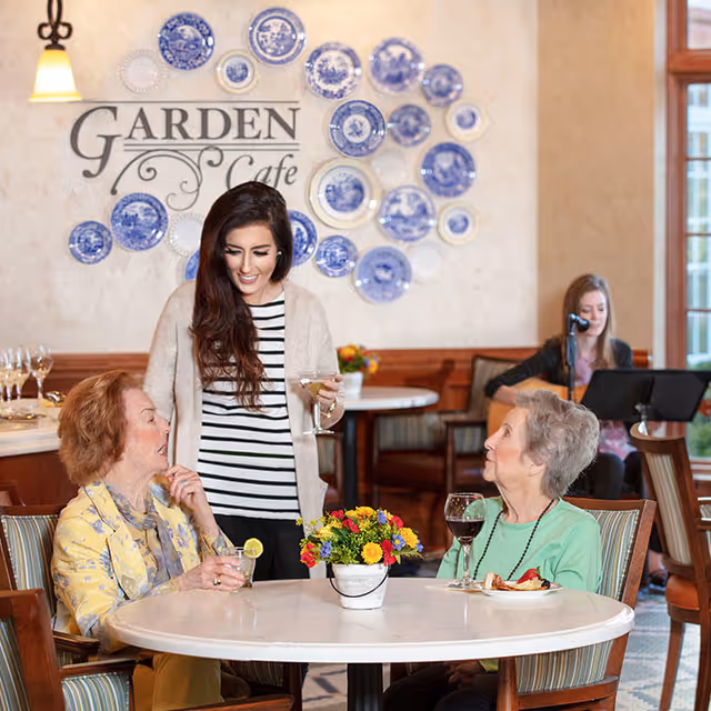 Two elderly women sitting at a round table with drinks and a plate of food, engaged in conversation with a younger woman standing beside them holding a drink. In the background, a musician is playing a guitar and singing. The wall behind them is decorated with blue and white plates and a sign that reads 'Garden Cafe'.