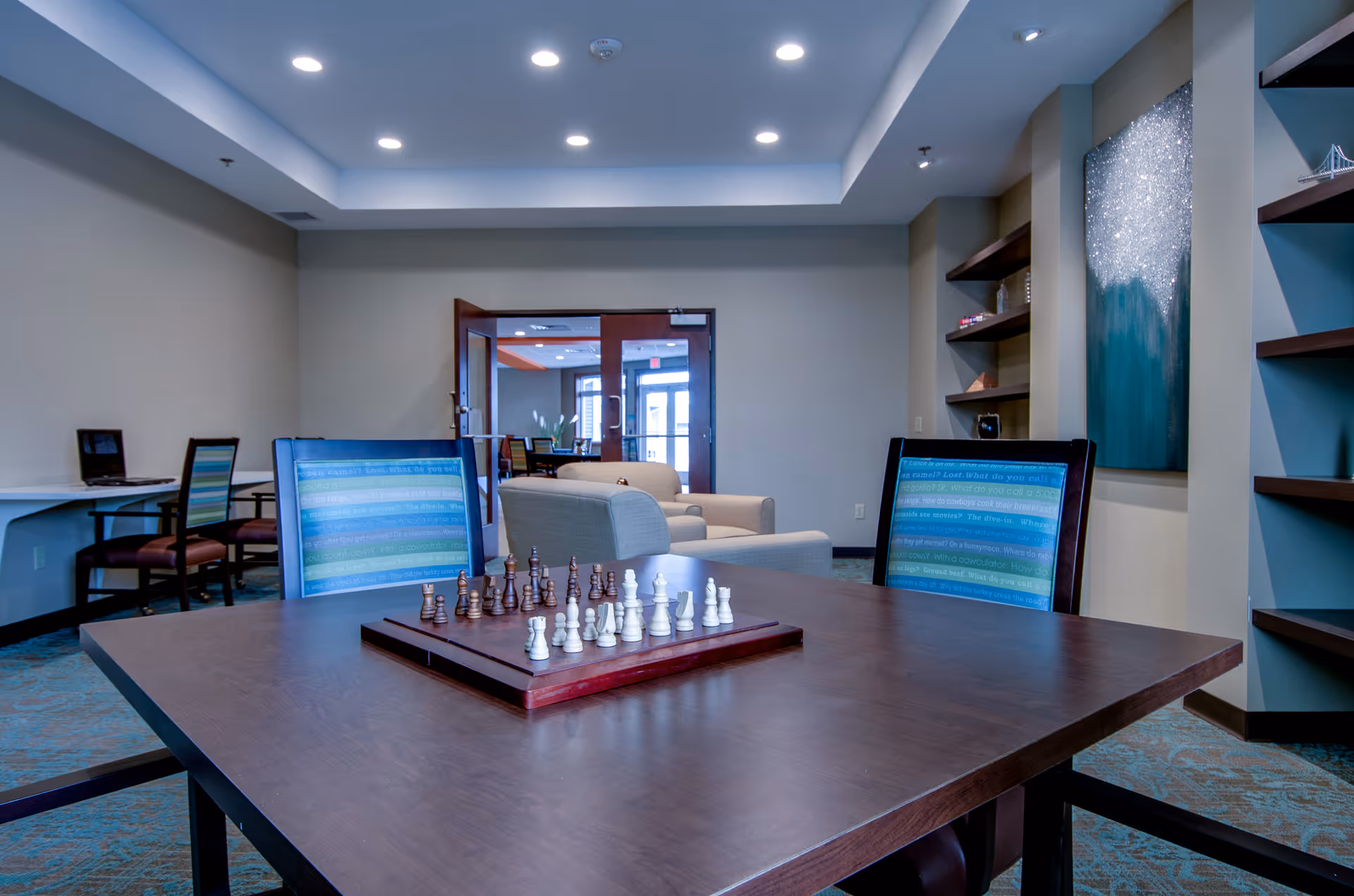 A well-lit common area in a senior living facility featuring a wooden table with a chessboard and chess pieces set up. Two chairs with blue and green striped upholstery are positioned at the table. In the background, there are beige armchairs, built-in shelves with decorative items, a large abstract painting on the wall, and a doorway leading to another room with more seating and natural light.