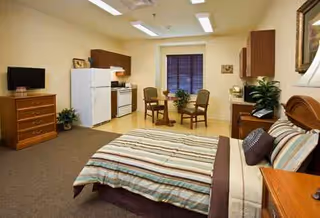 Interior studio apartment in a senior facility with a bed, kitchenette, and dining table.