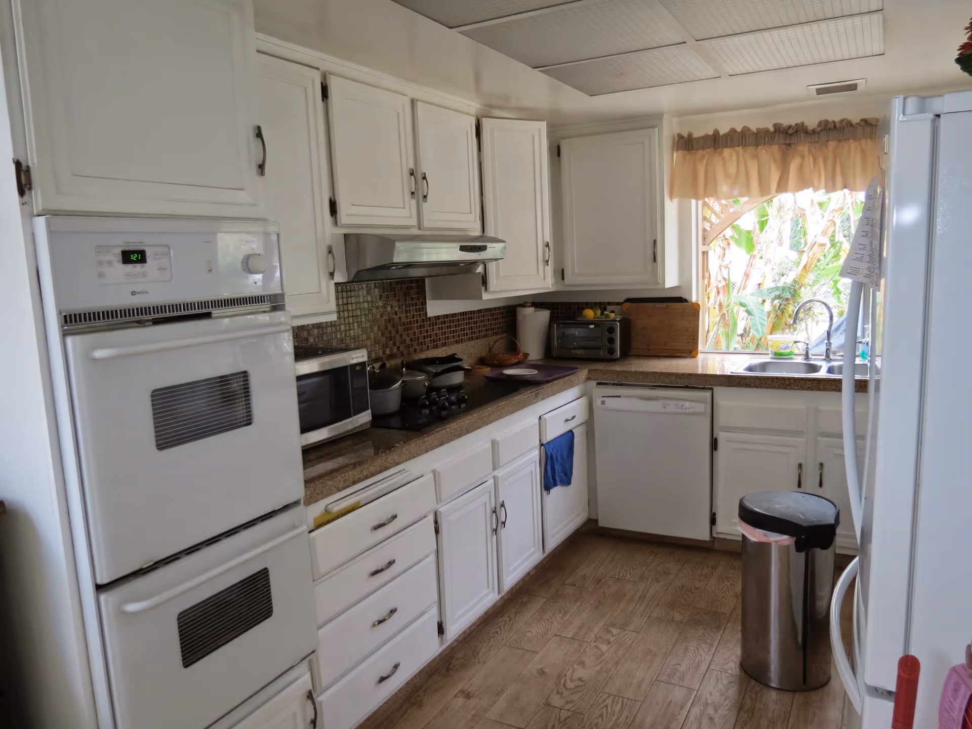 A kitchen with white cabinets and appliances, including a double oven, microwave, stove, dishwasher, and refrigerator. There is a window above the sink with a beige valance, and a trash can is placed near the refrigerator. The floor has a wood-like finish.