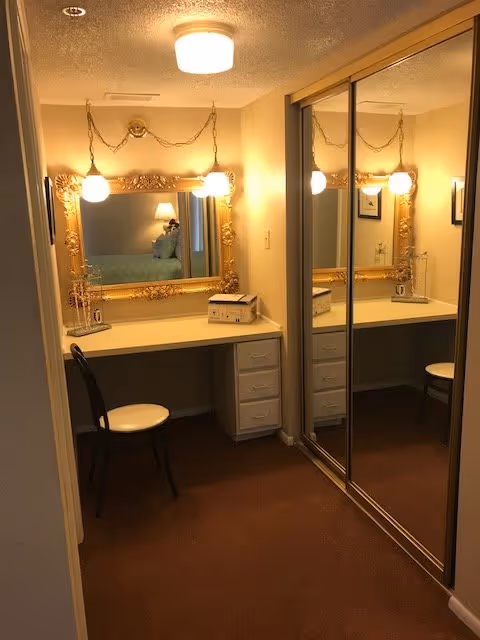 A small vanity area with a white desk and drawers, a black chair with a white seat cushion, and a large ornate gold-framed mirror hanging on the wall. Two hanging pendant lights with round bulbs illuminate the space. To the right, there is a large mirrored sliding closet door reflecting the vanity area. The room has beige walls and brown carpet.