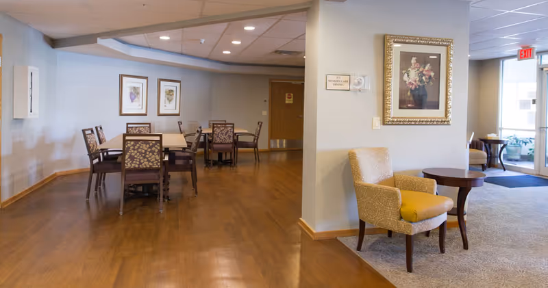An open dining and seating area with wooden floors, multiple tables and patterned chairs, and a small lounge chair and side table near the entryway.