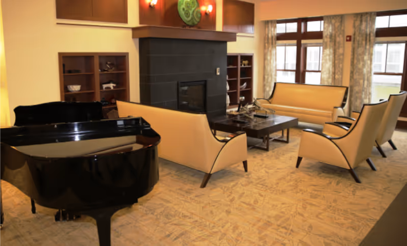 A bright communal lounge with beige sofas and armchairs arranged around a coffee table and fireplace, and a black grand piano by large windows.