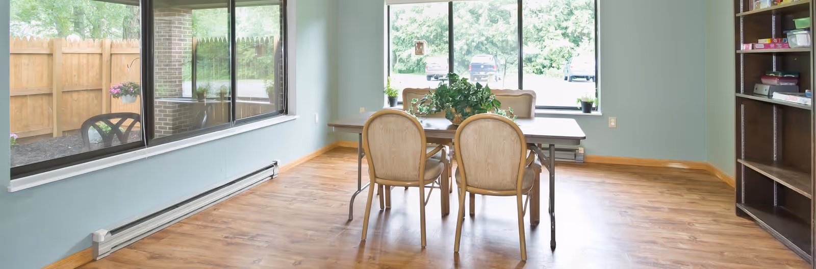 A bright room with large windows showing an outdoor fenced area. Inside, there is a rectangular table with four chairs around it and a green plant centerpiece. The floor is wooden, and there is a bookshelf on the right side of the room.