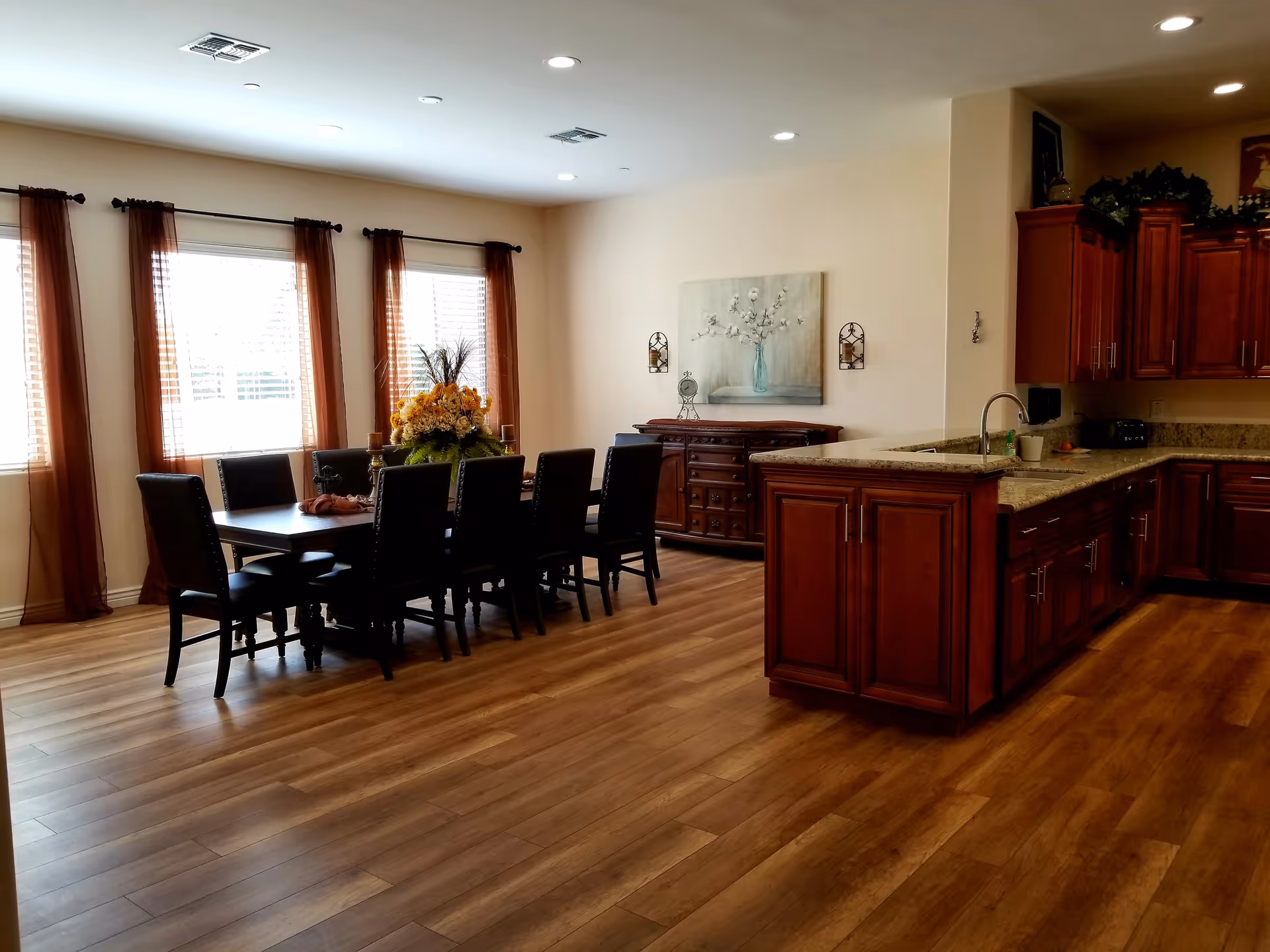 A spacious dining area with a large wooden dining table surrounded by eight dark chairs. The room has three windows with sheer brown curtains allowing natural light to fill the space. Adjacent to the dining area is a kitchen with dark wooden cabinets and granite countertops. The floor is made of light brown wood, and there is a decorative floral arrangement on the dining table.