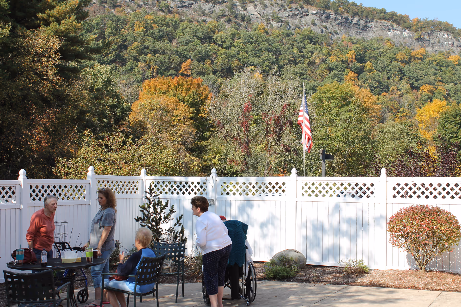 A group of elderly people and a caregiver are gathered outdoors in a fenced patio area with chairs and a table. Behind the white fence, there are trees with autumn foliage and a rocky hill in the background. An American flag is visible near the fence.