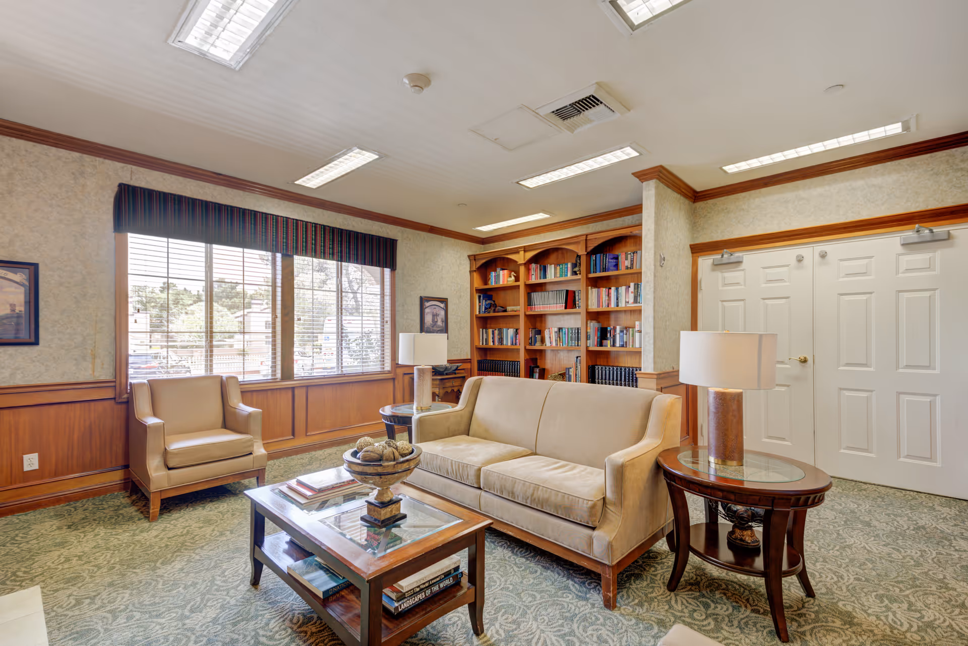 A cozy living room area in a senior living facility with a beige sofa, matching armchair, wooden coffee table with decorative items, side table with a lamp, large window with blinds, and a built-in wooden bookshelf filled with books.