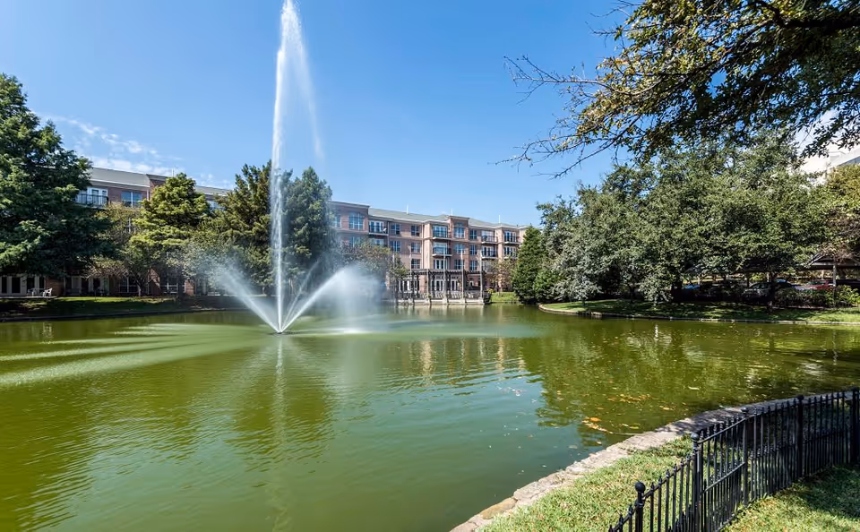 A large pond with a tall water fountain in the center, surrounded by trees and a black metal fence. In the background, there is a multi-story brick building under a clear blue sky.