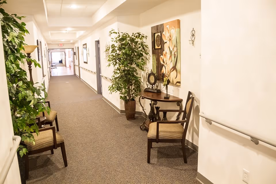 A well-lit hallway in a senior living facility with beige walls and carpeted floor. The hallway features handrails on both sides, several chairs with striped cushions, potted plants, a small decorative table with a clock and a flower vase, and framed artwork on the walls. The hallway extends into the distance with doors along the sides and an exit sign visible at the far end.