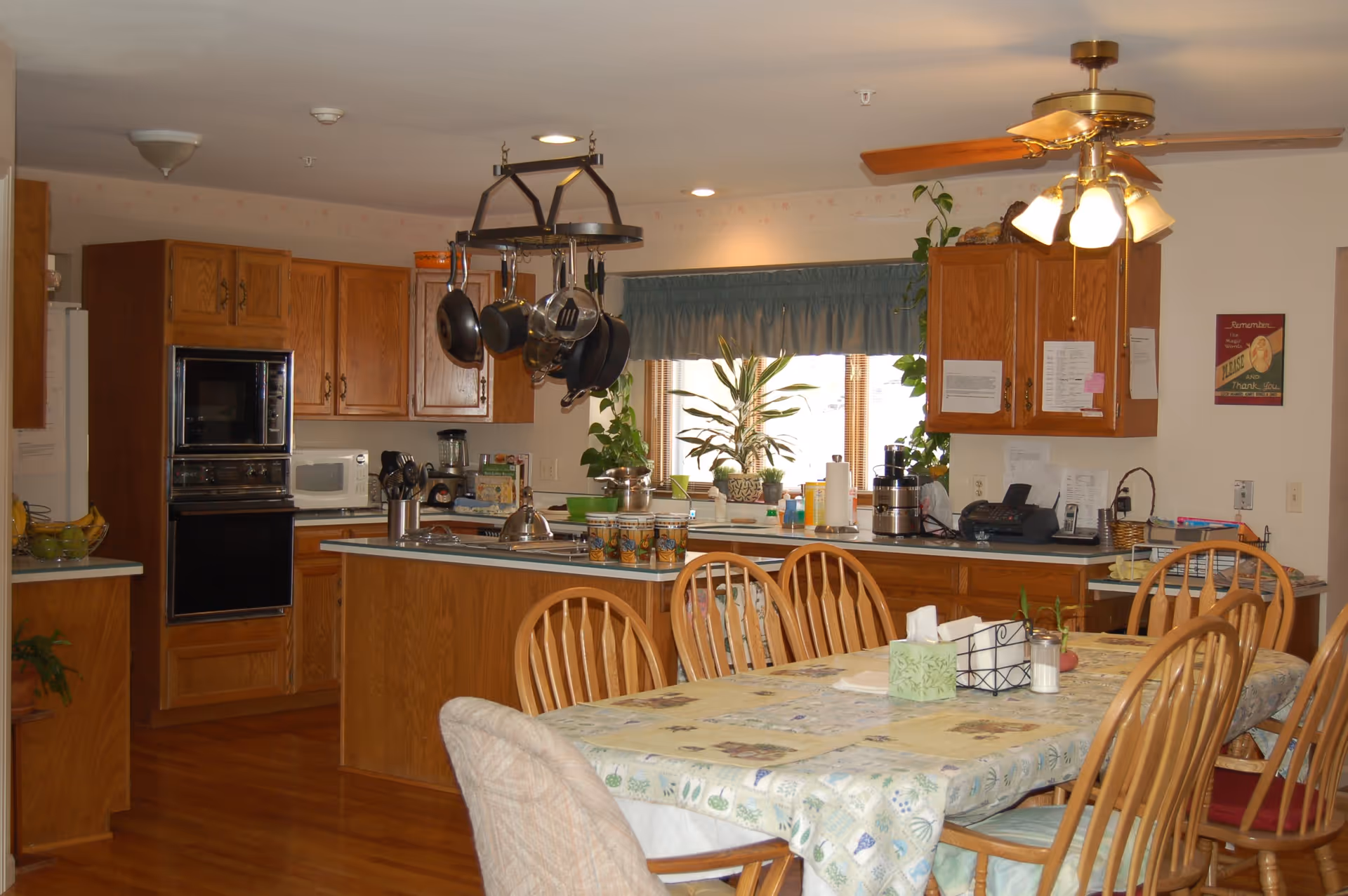 A warm and inviting kitchen and dining area with wooden cabinets and hardwood floors. The kitchen features a hanging rack with pots and pans, a microwave, oven, and various kitchen appliances on the counters. The dining table is covered with a patterned tablecloth and surrounded by wooden chairs. A ceiling fan with lights hangs above the dining area, and a window with a blue valance lets in natural light, with several potted plants on the windowsill.