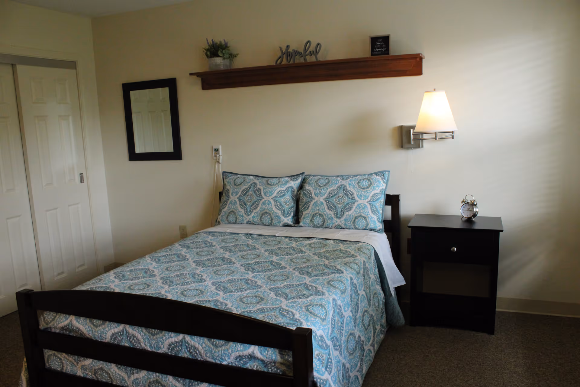 A simple furnished bedroom with a bed dressed in blue patterned bedding, a nightstand with an alarm clock, a wall-mounted lamp, and a shelf above the headboard.