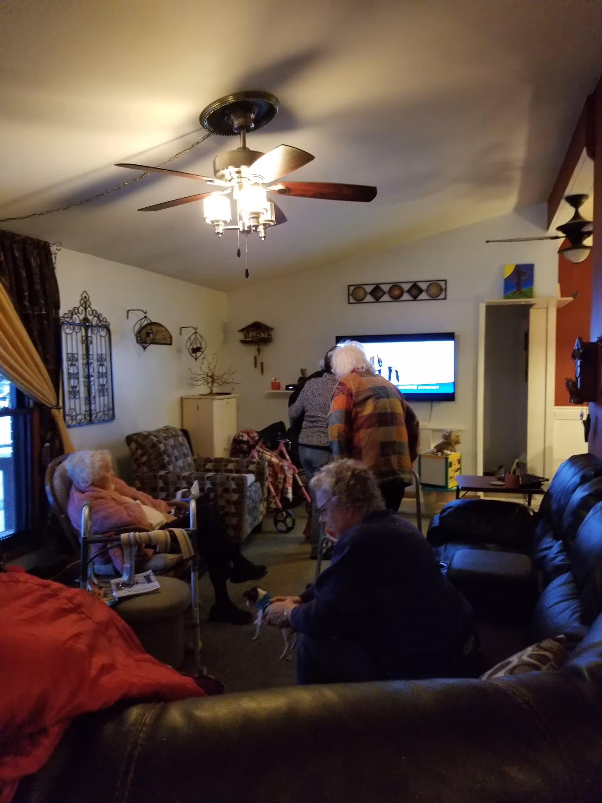 A cozy living room with several elderly people sitting and interacting. There are armchairs and a leather couch arranged around the room. A ceiling fan with lights is on, and a flat-screen TV is mounted on the wall. The room has warm lighting and various decorations on the walls.