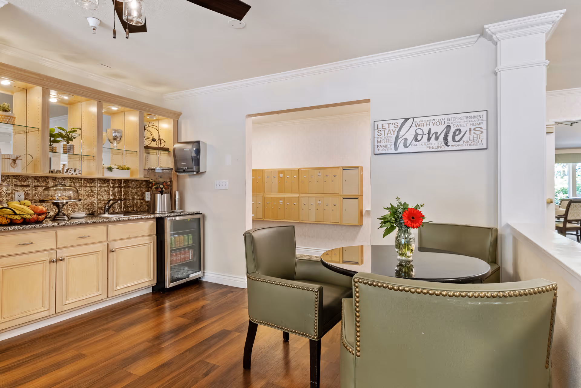 A cozy interior space with a small round table and three green upholstered chairs. On the table is a glass vase with a red flower and greenery. To the left is a countertop with a fruit basket, a cake stand, a water dispenser, and a small refrigerator stocked with beverages. Above the countertop are shelves with decorative items and lighting. In the background, there are multiple mailboxes mounted on the wall and a wall sign that reads 'home' with additional text.