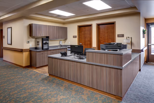 Reception/nurse station with a desk, computers, printer, cabinets, and two closed wooden doors in a hallway.