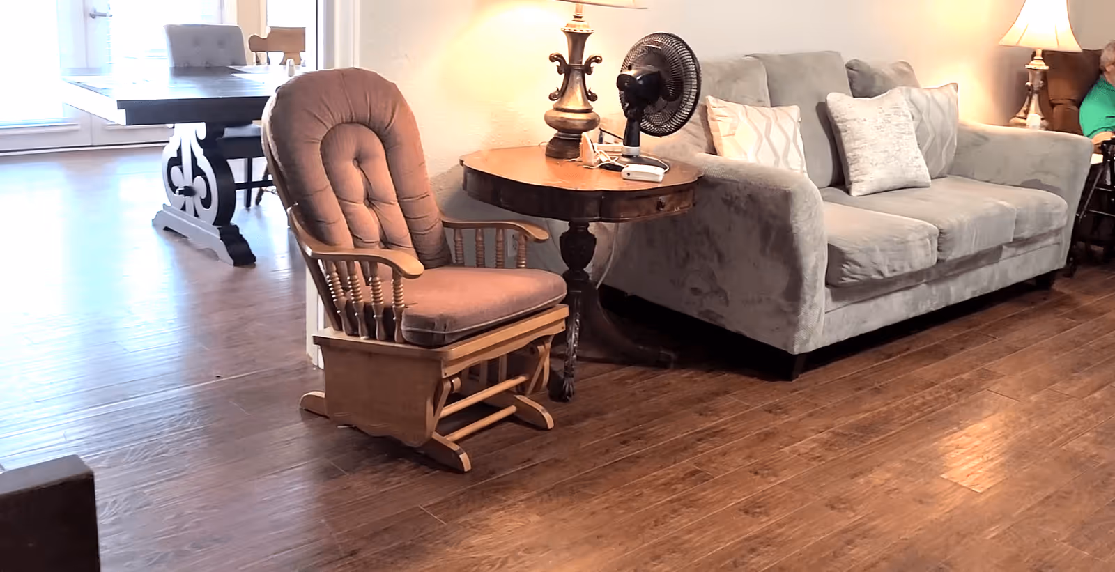 A cozy living room area featuring a cushioned wooden rocking chair, a round wooden side table with a lamp and a small fan on it, and a gray upholstered sofa with decorative pillows. In the background, there is a dining table with chairs and a person sitting in a wheelchair near another lamp.