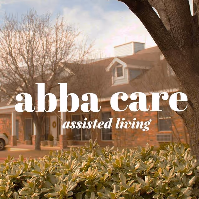 Exterior view of a brick assisted living facility with a tree and green bushes in the foreground under a partly cloudy sky.