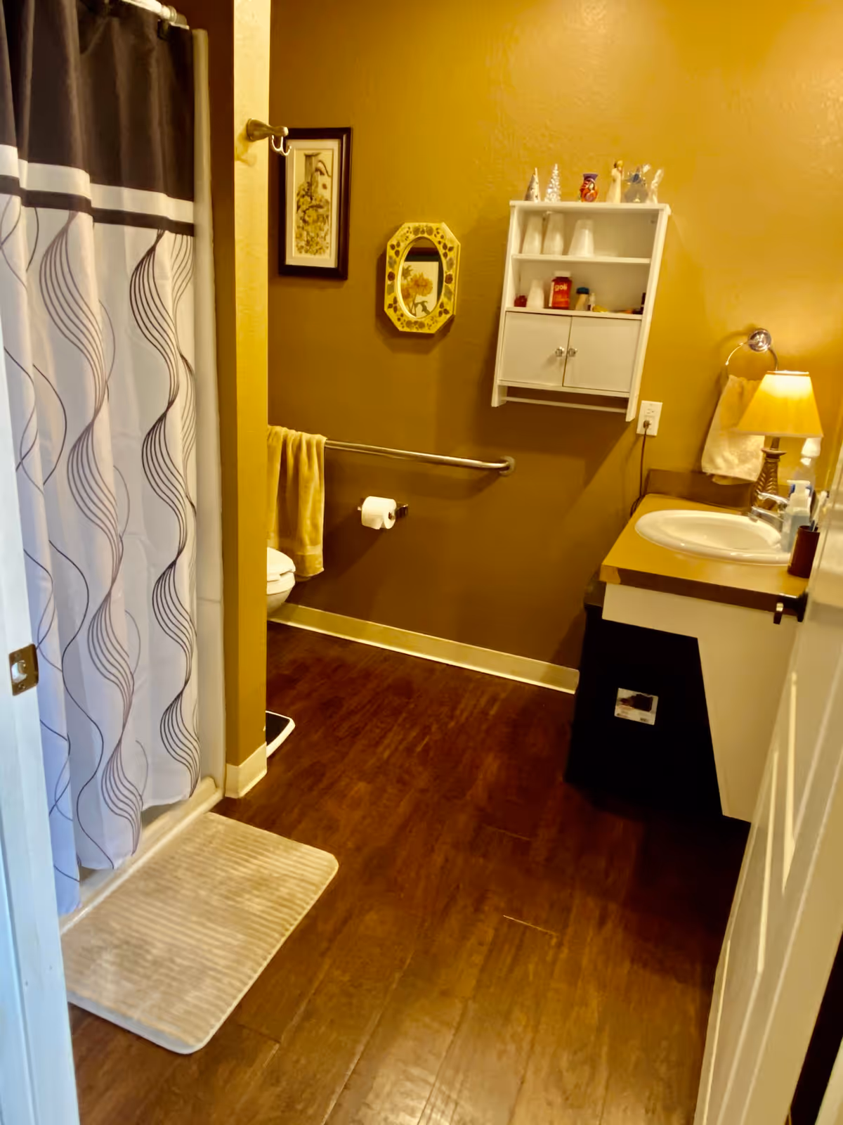 A bathroom with brown walls and wooden floor. There is a white shower curtain with a wavy pattern on the left, a white bath mat on the floor, a toilet with a yellow towel hanging on a grab bar, a white wall cabinet with cups and small decorative items, a sink with a countertop, a lamp with a yellow shade, and a towel hanging on a ring near the sink. Two framed pictures hang on the wall.