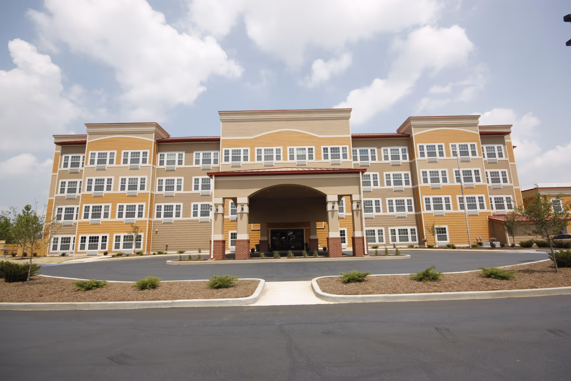 Front exterior of a four-story yellow and beige senior living building with a covered entrance and circular driveway under a partly cloudy sky.