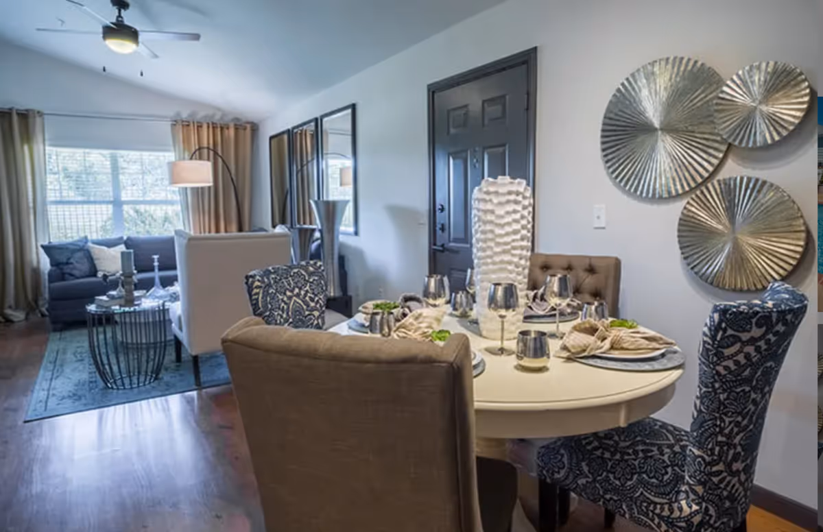 A cozy living and dining area with a round dining table set for four, featuring silverware and napkins. The dining chairs are upholstered in patterned and plain fabrics. Behind the dining area is a living room with a sofa, armchair, coffee table, floor lamp, and large windows with curtains. Decorative metallic wall art is displayed on the wall near the dining table.