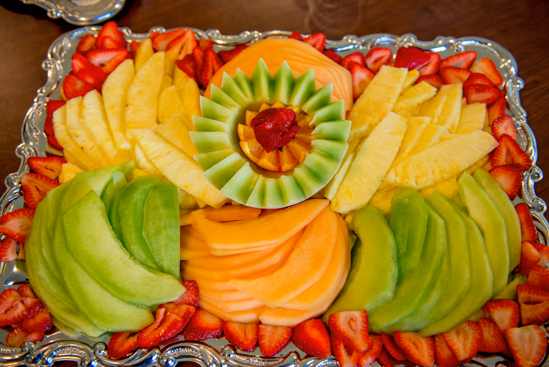 Decorative silver platter with arranged slices of honeydew, cantaloupe, pineapple, and strawberries.