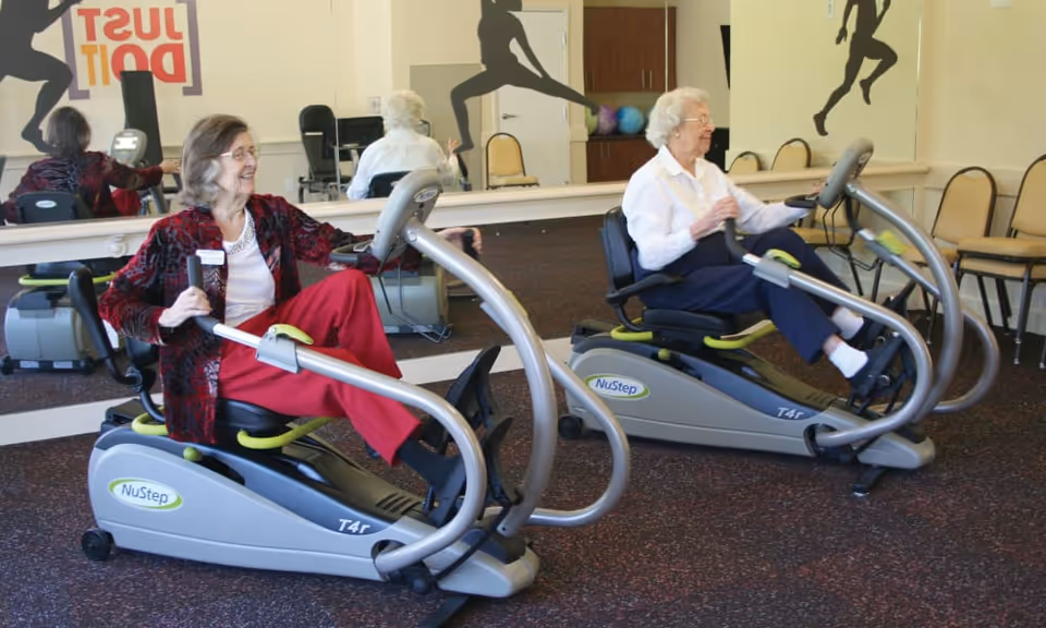 Two elderly women exercising on NuStep T4r recumbent cross trainers in a fitness room with a large mirror and silhouettes of people exercising on the wall.