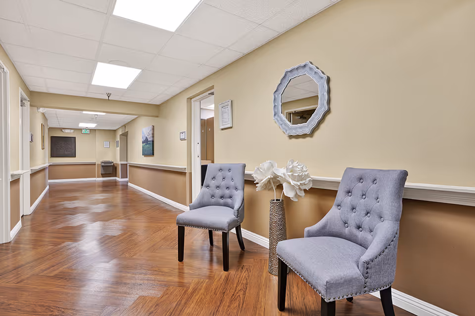 A clean and well-lit hallway in a senior living facility with wood-patterned flooring and beige walls. Two gray upholstered chairs with button tufting are placed against the right wall, next to a tall vase with white decorative flowers. A decorative mirror hangs above the chairs. The hallway extends into the distance with doors and framed artwork on the walls.